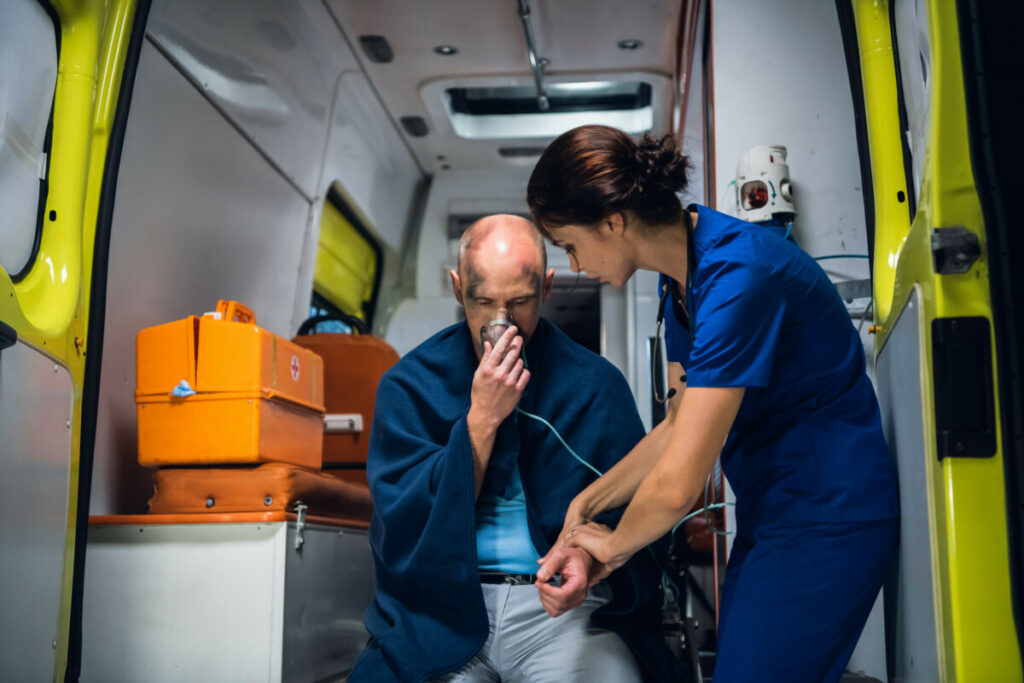 Man sits with oxygen mask, woman in medical uniform holds his hand