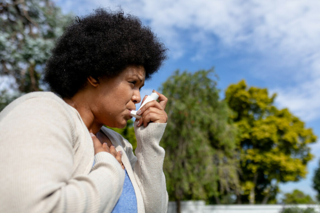 African american mid adult woman with hand on chest using asthma inhaler while standing outdoors. unaltered, healthcare, asthmatic, reliever asthma inhaler, inhaling and illness concept.